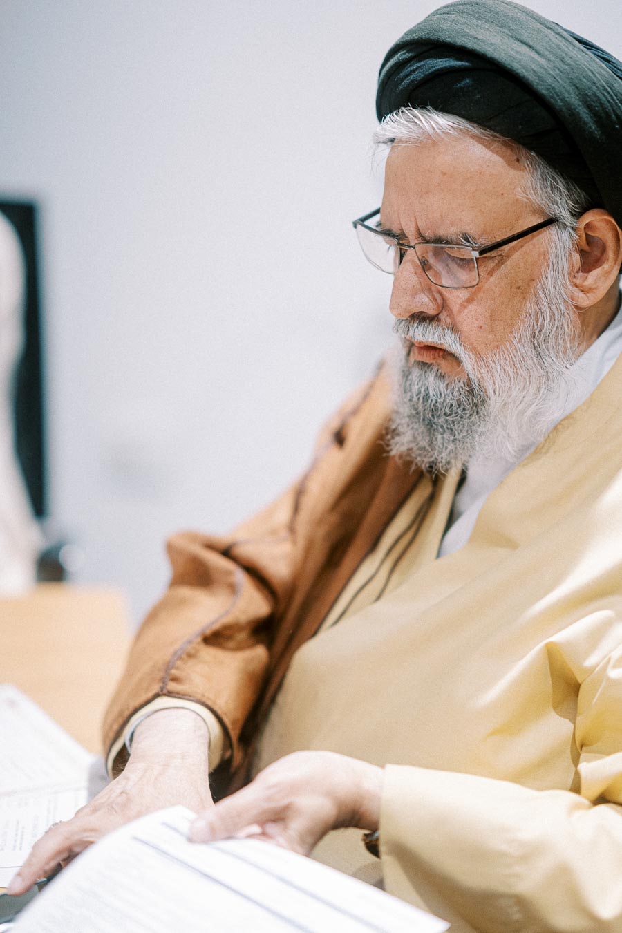 Elderly man wearing traditional attire and reading documents at a desk, focused and deep in thought.