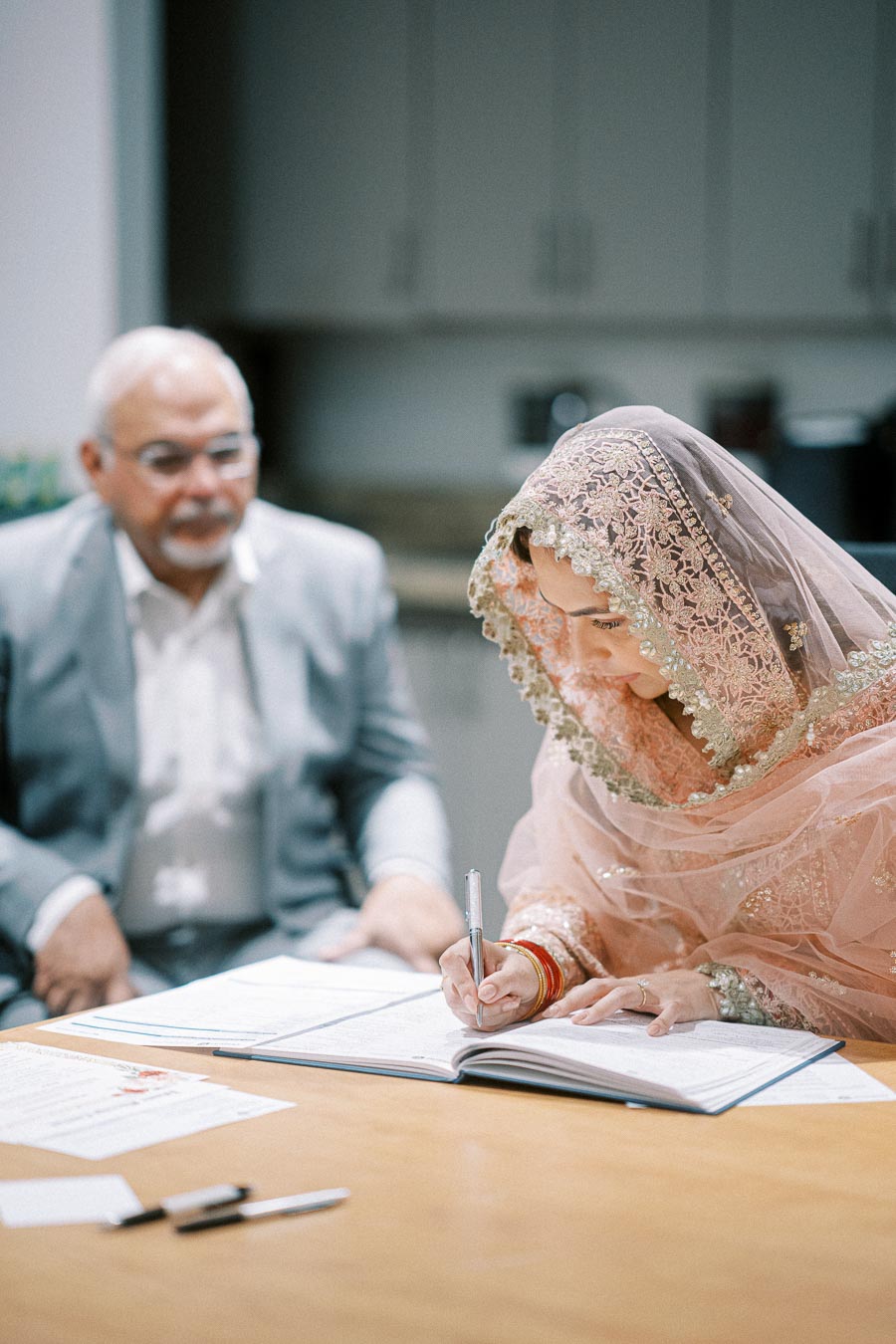 A bride in a pink embroidered traditional outfit signs a marriage document as part of a wedding ceremony, with an elder man seated beside her.