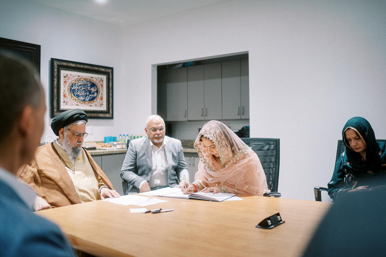 A group of people gathered around a table in a formal meeting setting, with one person in traditional attire signing a document. A framed artwork with calligraphy is visible in the background.