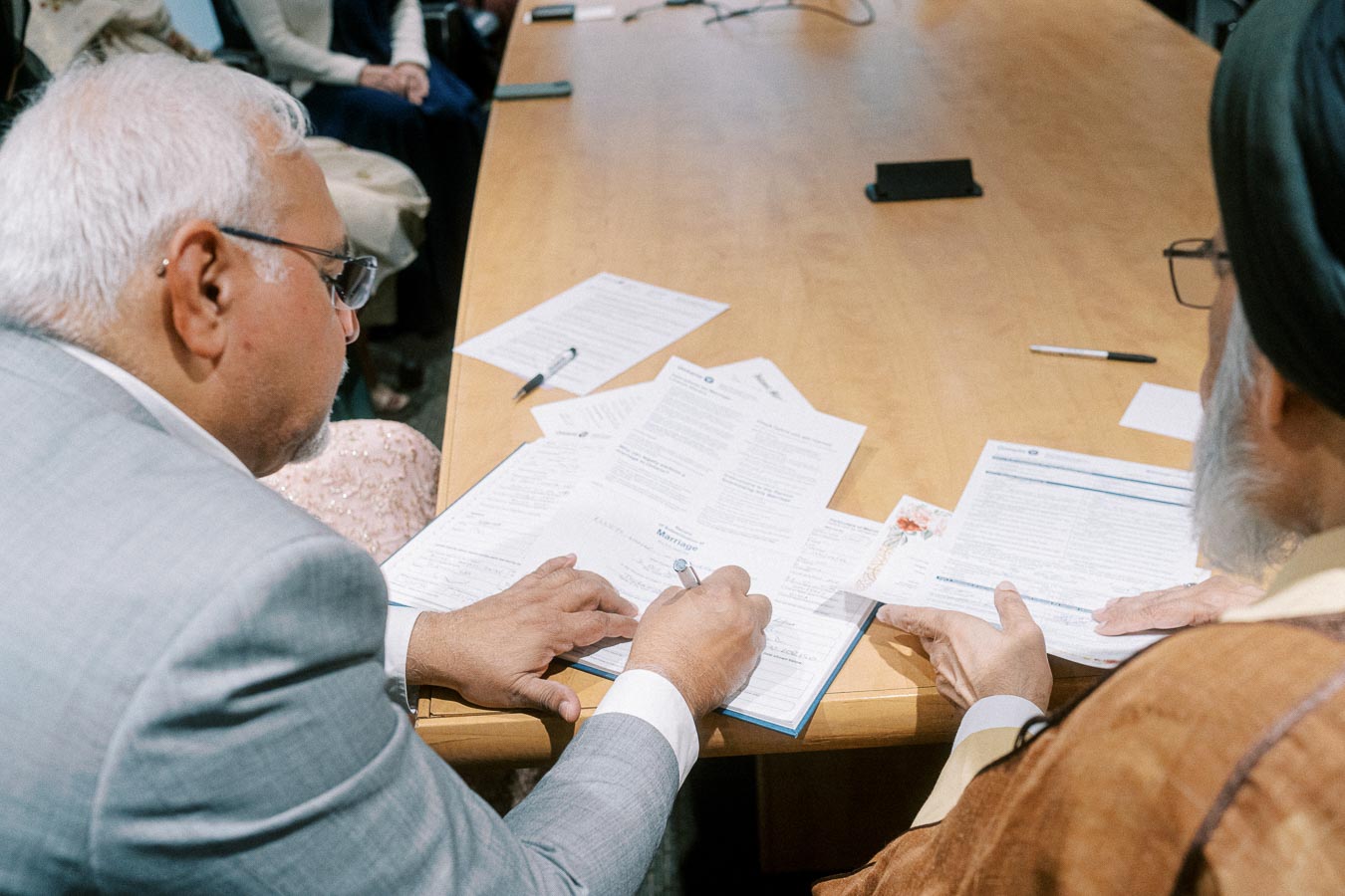 Two individuals reviewing and signing official documents at a conference table, with paperwork and pens visible, indicating a formal meeting or agreement process.