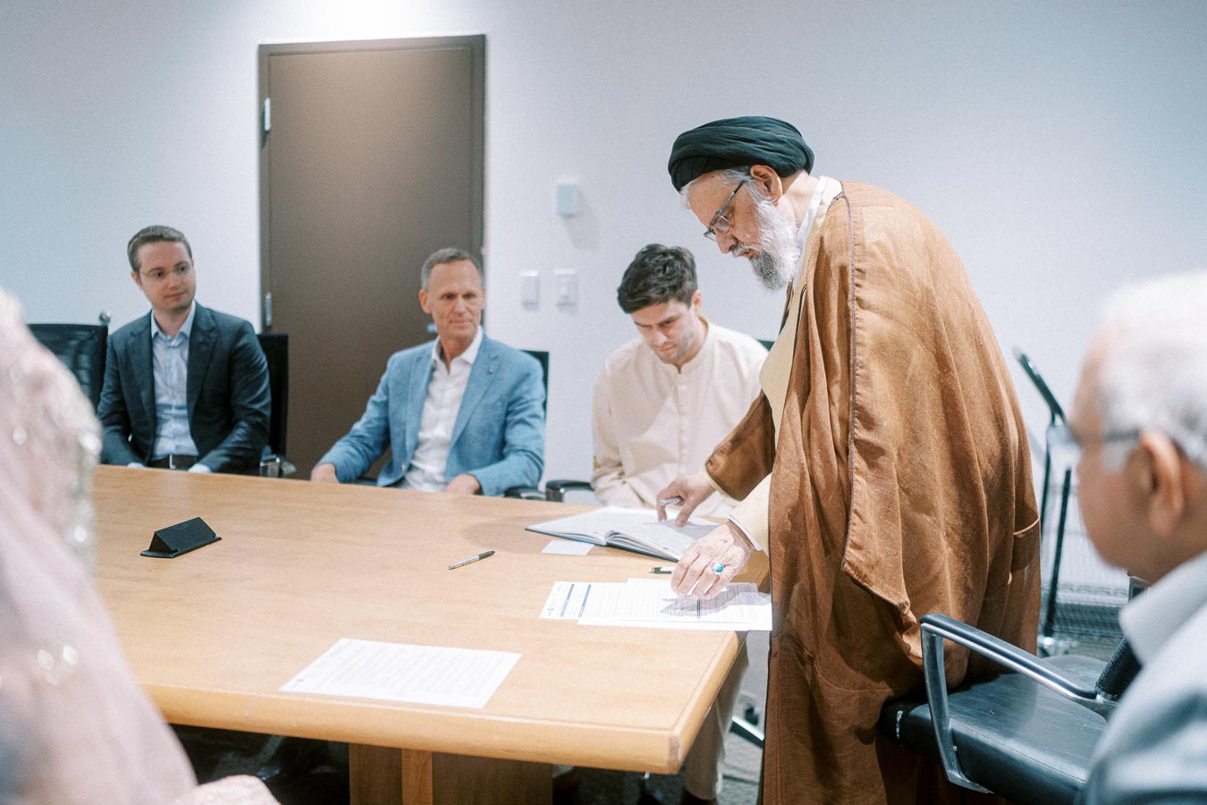 A group of men in a business meeting, gathered around a conference table. One man, wearing traditional attire, is standing and reviewing a document, while others are seated attentively, suggesting a discussion or negotiation setting.