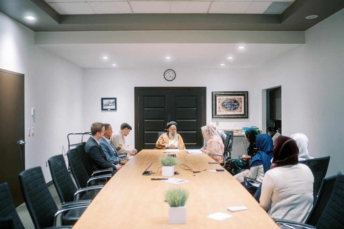 A multicultural group of individuals in a formal meeting room, centered around a large wooden table with decorative elements and artwork on the walls, signifying a diverse and inclusive discussion setting.