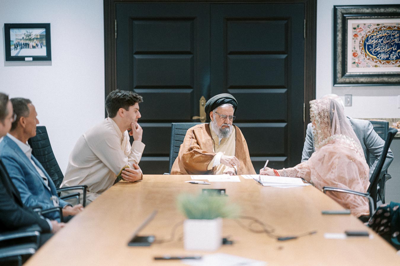 Meeting in a conference room with diverse individuals, including a person in traditional attire, discussing documents at a long table.
