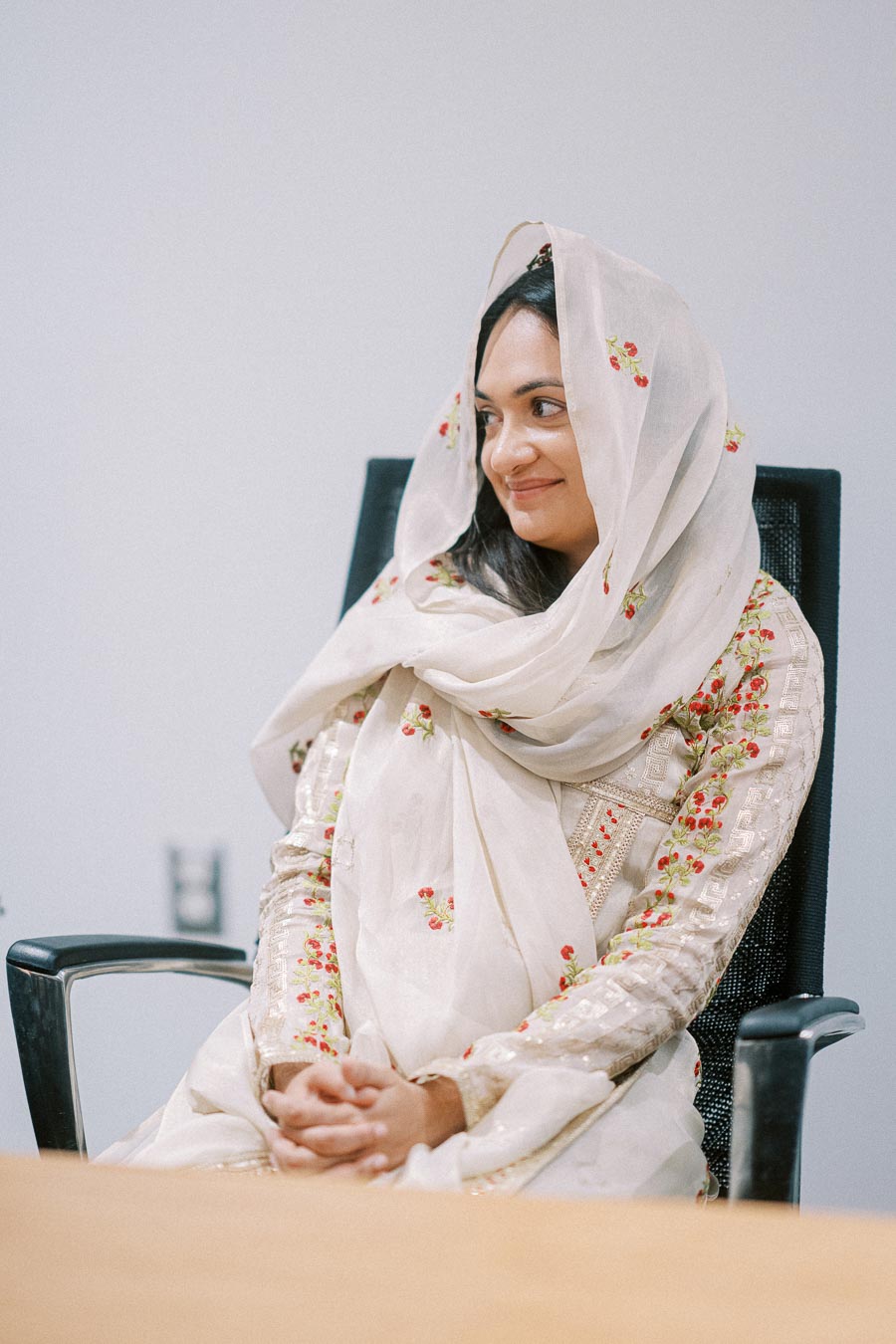 Woman wearing a traditional embroidered outfit with a scarf, seated on a chair, smiling and looking to the side in a bright room.