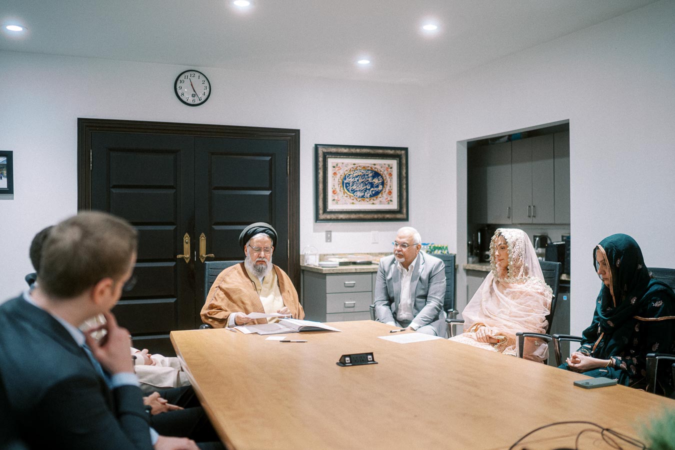 A group of people in traditional and formal attire sitting around a conference table, engaged in a meeting in a well-lit office setting. A clock and framed artwork are visible on the wall in the background.