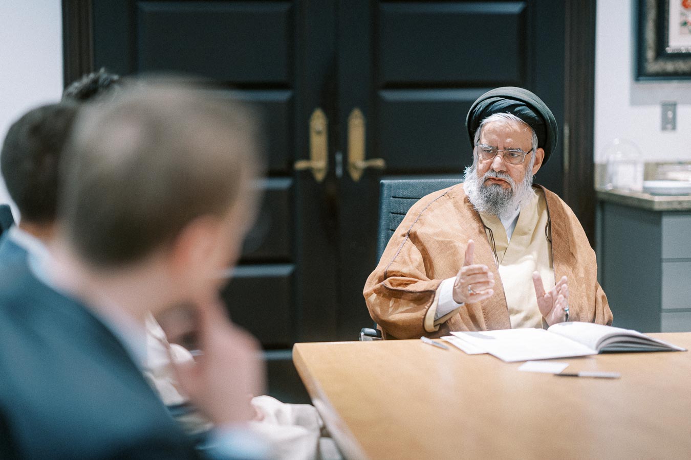 Elderly man in traditional attire speaking during a meeting with others at a conference table in an office setting.
