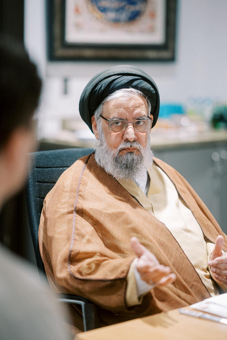 An elderly man in traditional attire engages in conversation in an office setting.