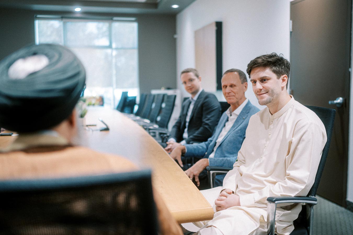 A group of people seated at a conference table in a meeting room, engaged in discussion. The room has a large window with natural lighting.