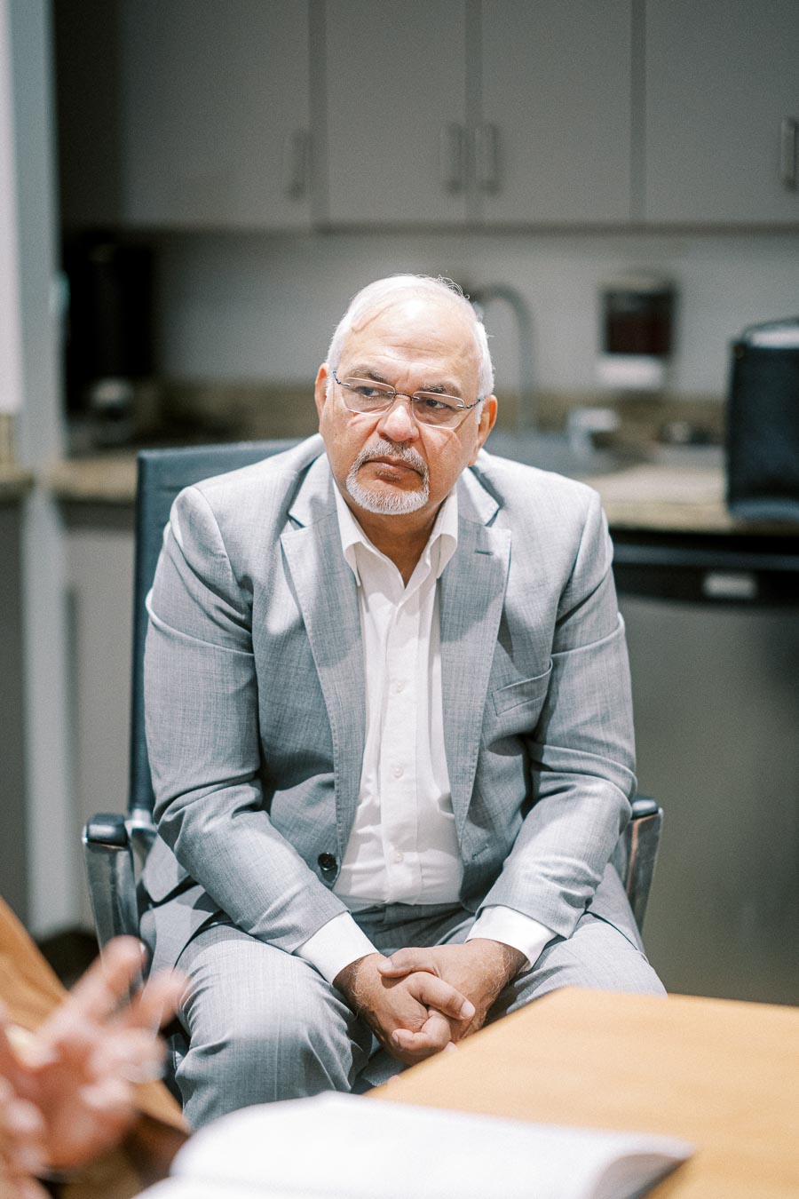 Elderly man in a gray suit sitting in an office meeting, looking attentive and engaged.