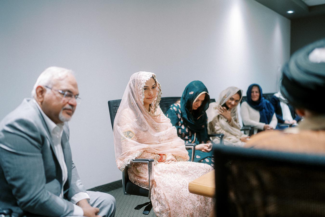 Group gathered in a conference room, featuring a woman in a traditional outfit with a delicate lace veil, sitting with others dressed in formal and cultural attire, engaged in conversation.