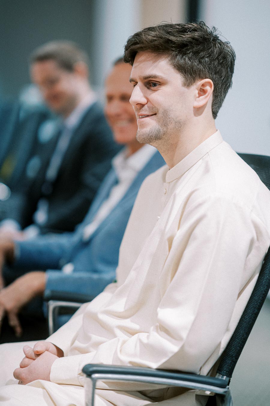 Young man in a white traditional outfit smiling during a professional meeting, sitting among suited colleagues in a modern office setting.