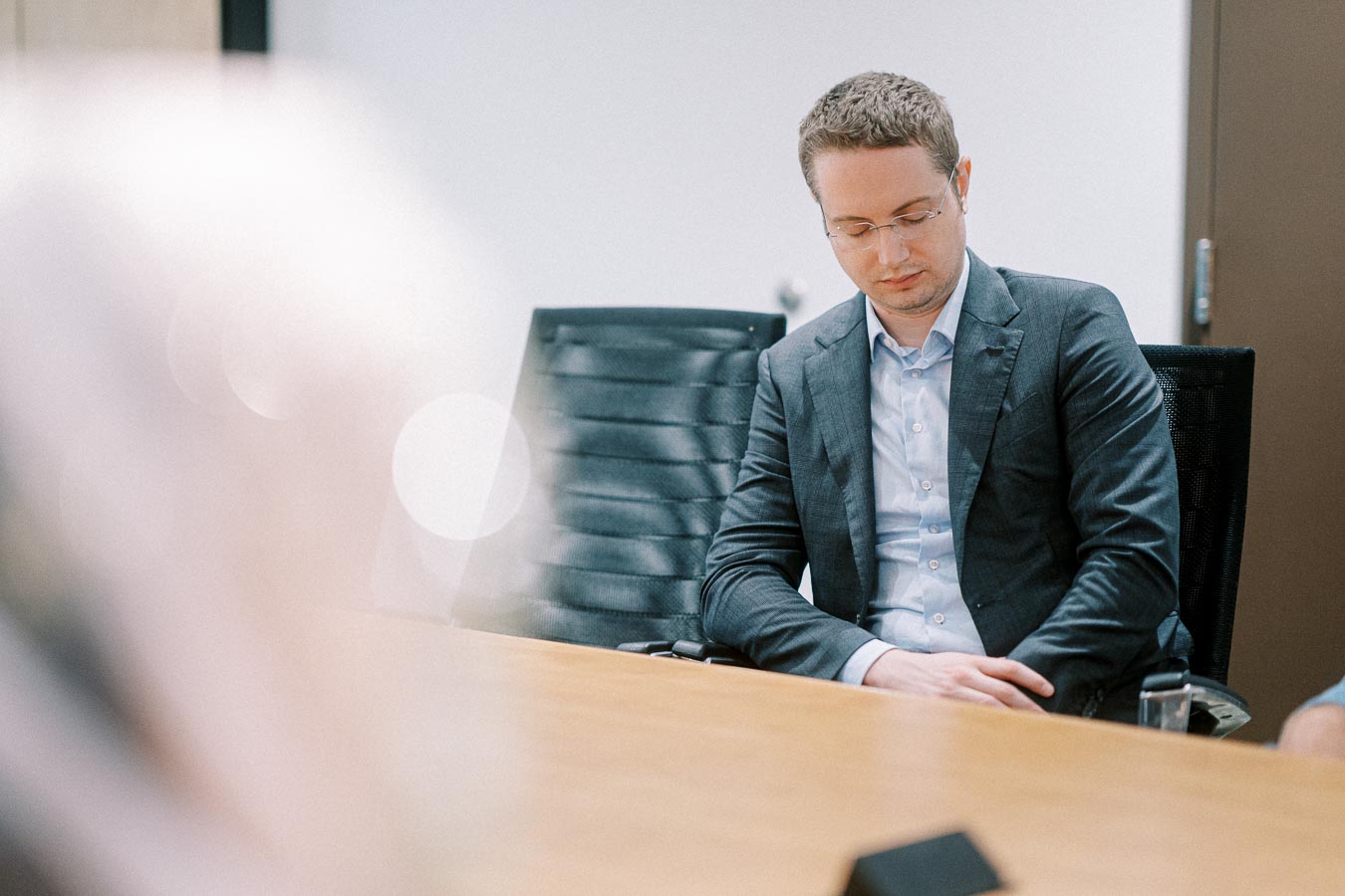 Male professional in a suit sitting thoughtfully in a conference room, resting his hands on a table, with soft lighting and a modern office chair, conveying a business meeting atmosphere.