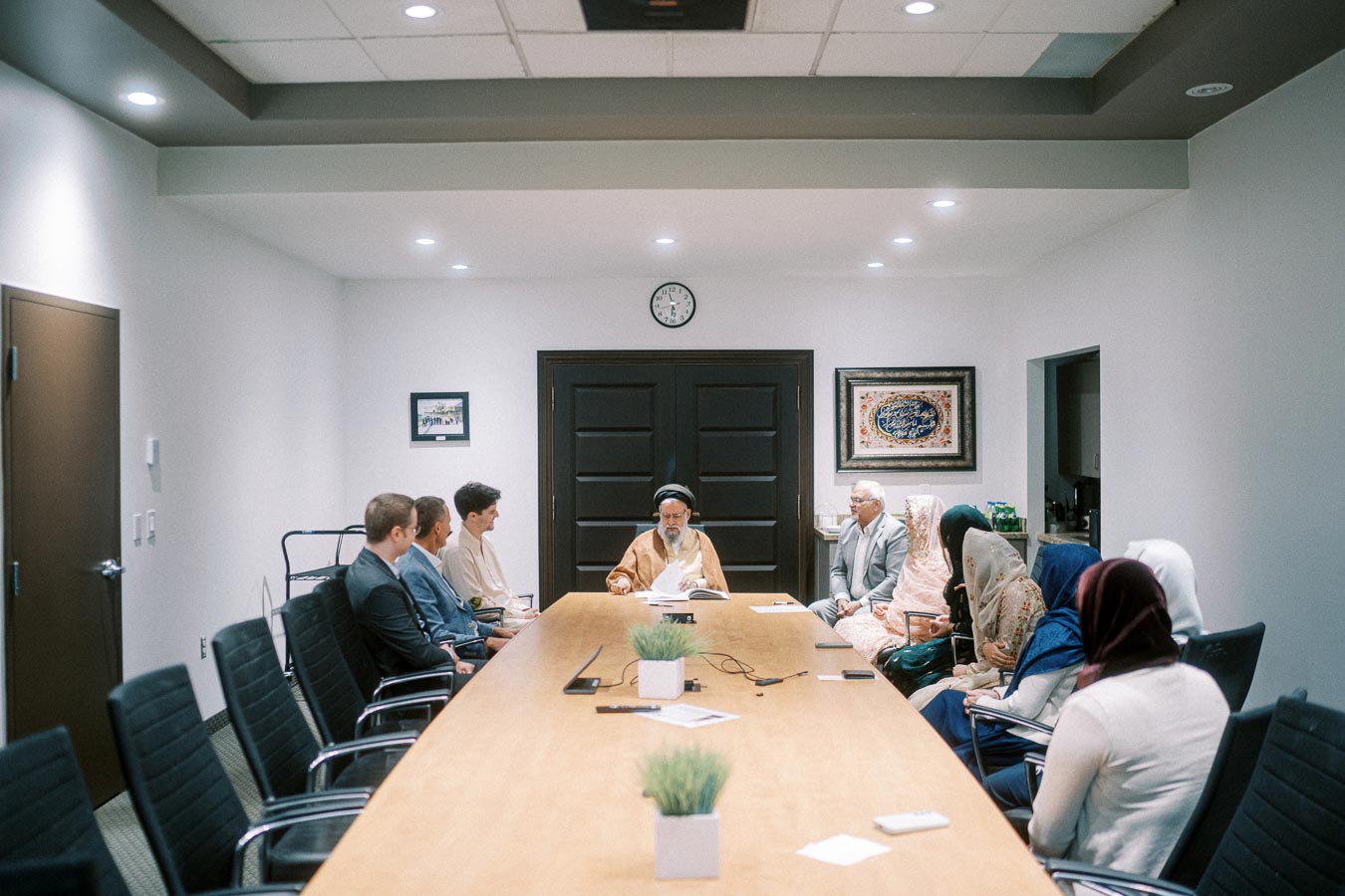 Meeting in a modern conference room with diverse participants seated around a long table, led by a figure in traditional attire at the head, under bright ceiling lights.