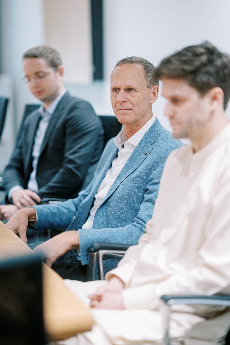 Three professionals in a business meeting, with a focus on a man in a blue blazer attentively listening.