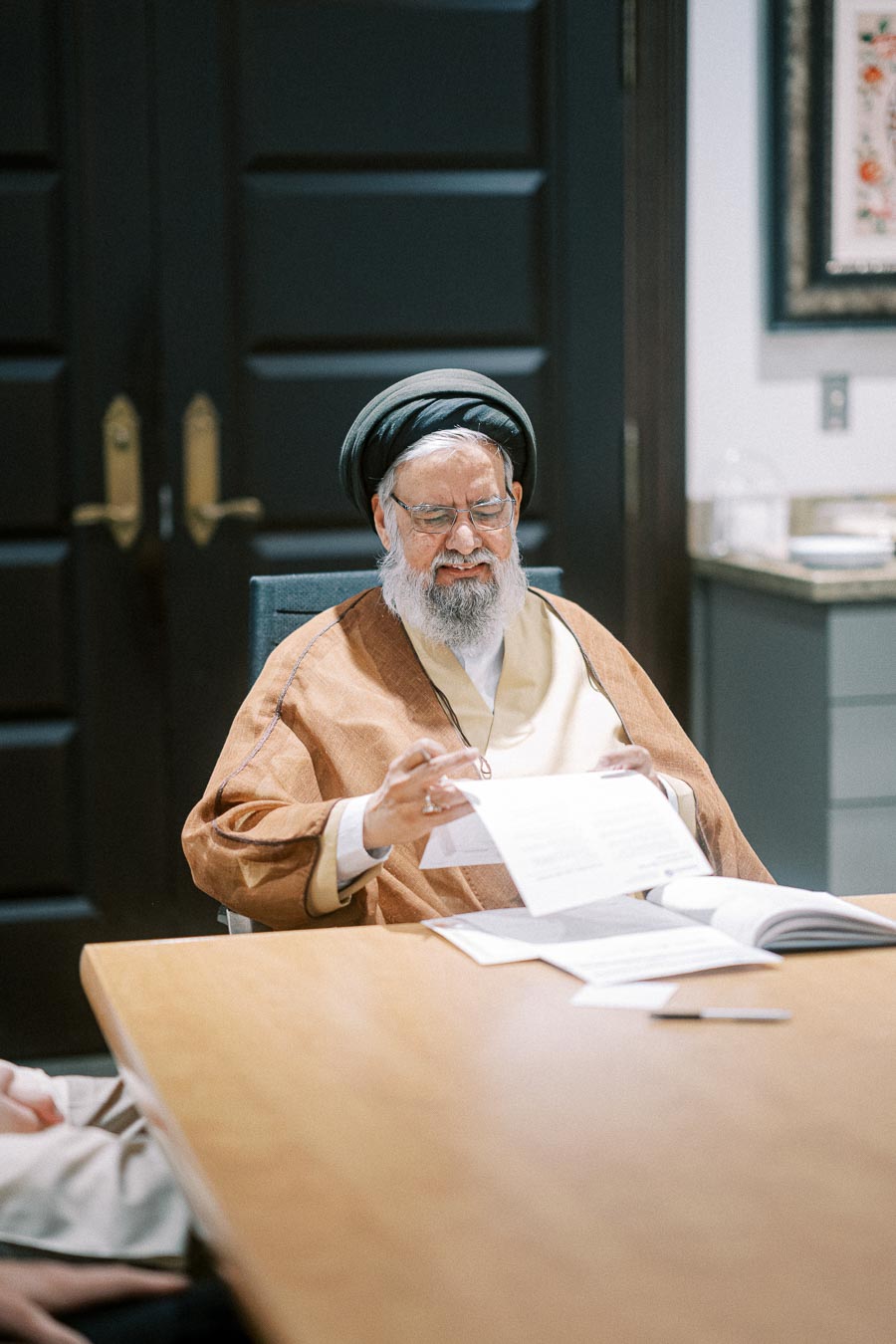 Elderly man in traditional attire examining documents at a wooden table in a well-lit office setting.