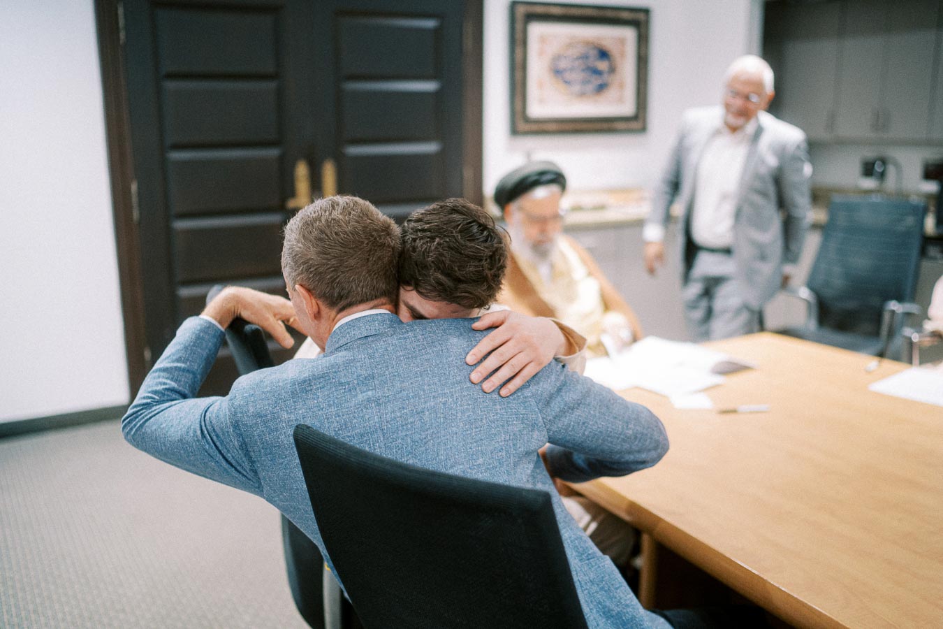 A comforting embrace between two men in a modern office setting, with colleagues discussing business at a conference table in the background.