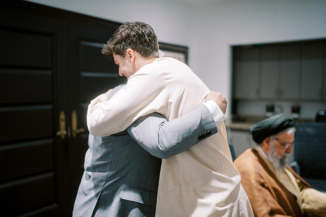 A man in a light sweater and another in a gray suit embrace warmly in an indoor setting, symbolizing friendship or reunion, while a seated man in traditional attire is visible in the background, focused on his own activity.