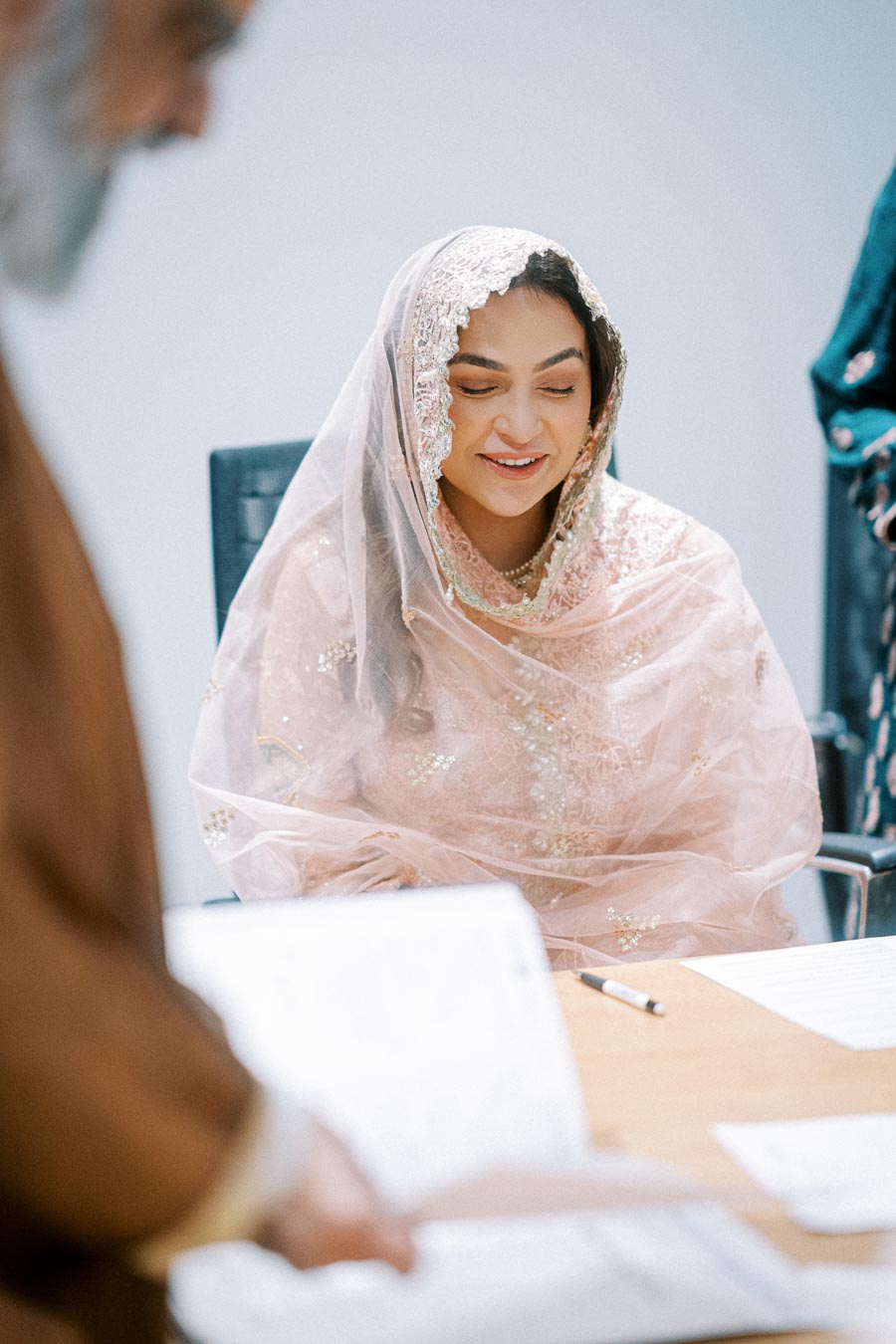 A woman in a pink traditional dress and veil smiling during a formal ceremony, seated at a table with documents and a pen, inside a bright room.
