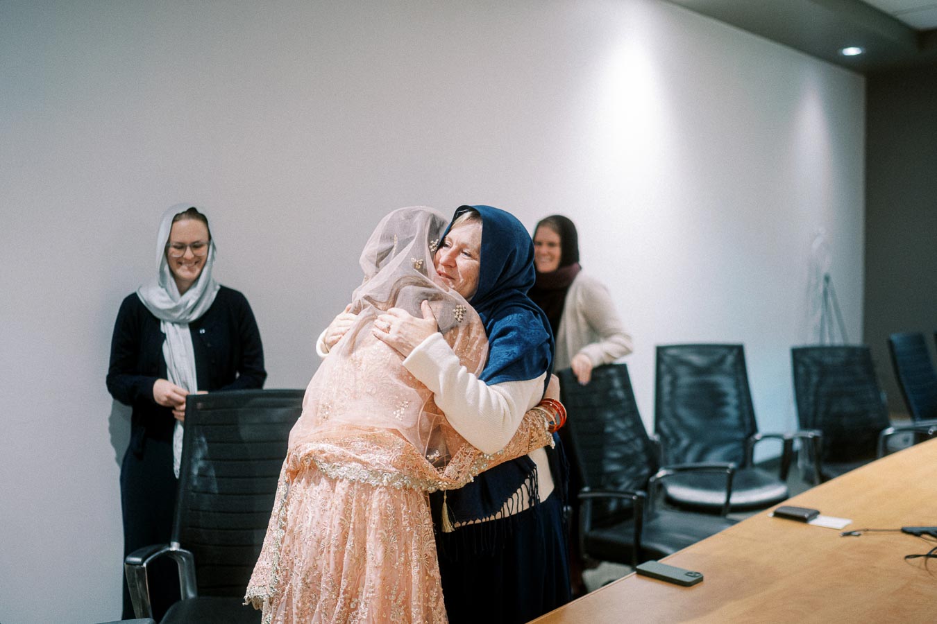 A group of women in a meeting room, two hugging warmly while others smile, creating a sense of community and connection, with chairs and a table in the background.