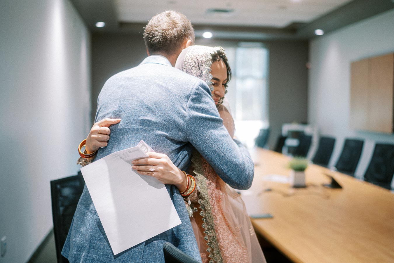 A couple embracing joyfully in a brightly lit conference room, with the man holding a document, possibly a marriage certificate, while the woman wears traditional attire and jewelry.