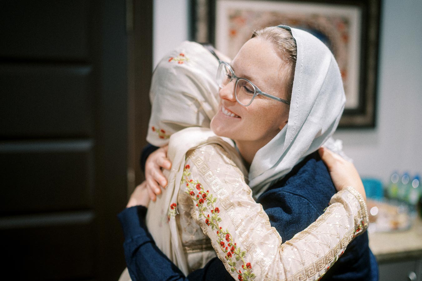 Two women wearing headscarves warmly embracing, one smiling broadly, in a culturally rich indoor setting.