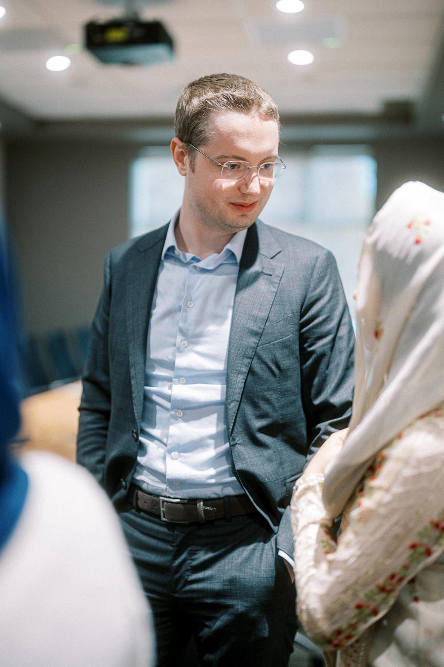 Man in a suit with glasses engaged in conversation at a social or business event, with a woman in traditional attire in the foreground, softly blurred.