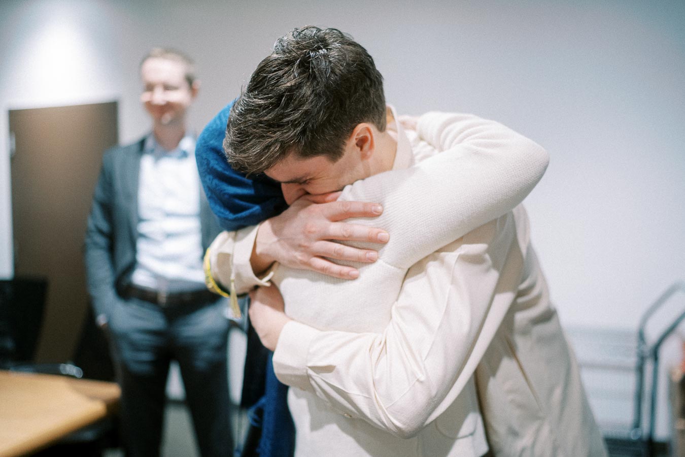 A person in a light sweater embraces another person warmly in an office setting, with a smiling man in a suit observing in the background.