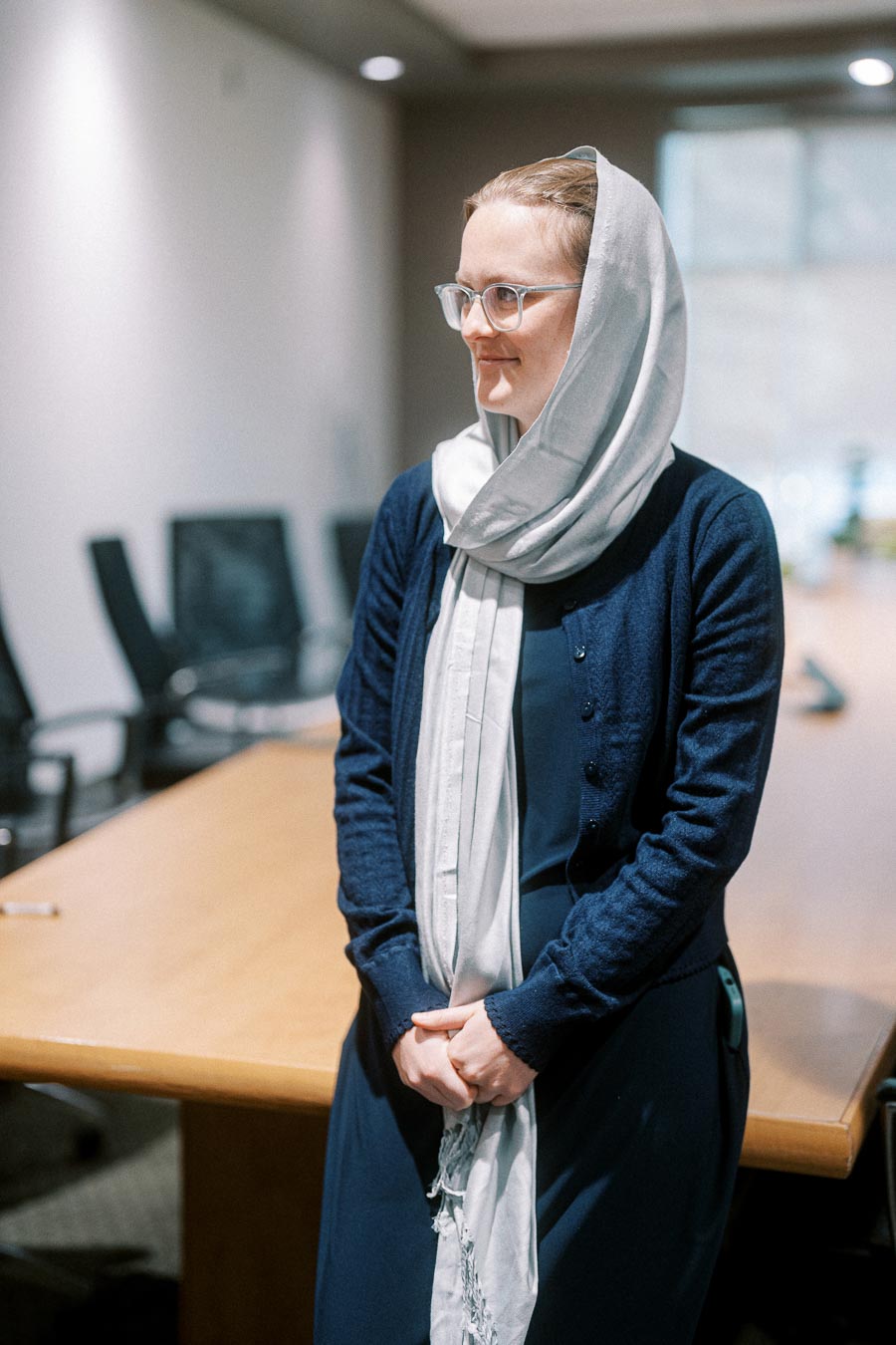 A person wearing a light gray headscarf and glasses stands in a conference room, dressed in a navy blue outfit, with a long wooden table and office chairs in the background, suggesting a professional setting.