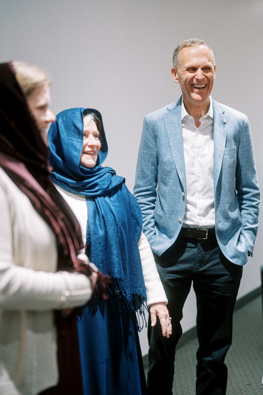 Three people smiling and engaging in a conversation, with one man in a light blue blazer and two women wearing headscarves, creating a warm and friendly atmosphere in an indoor setting.