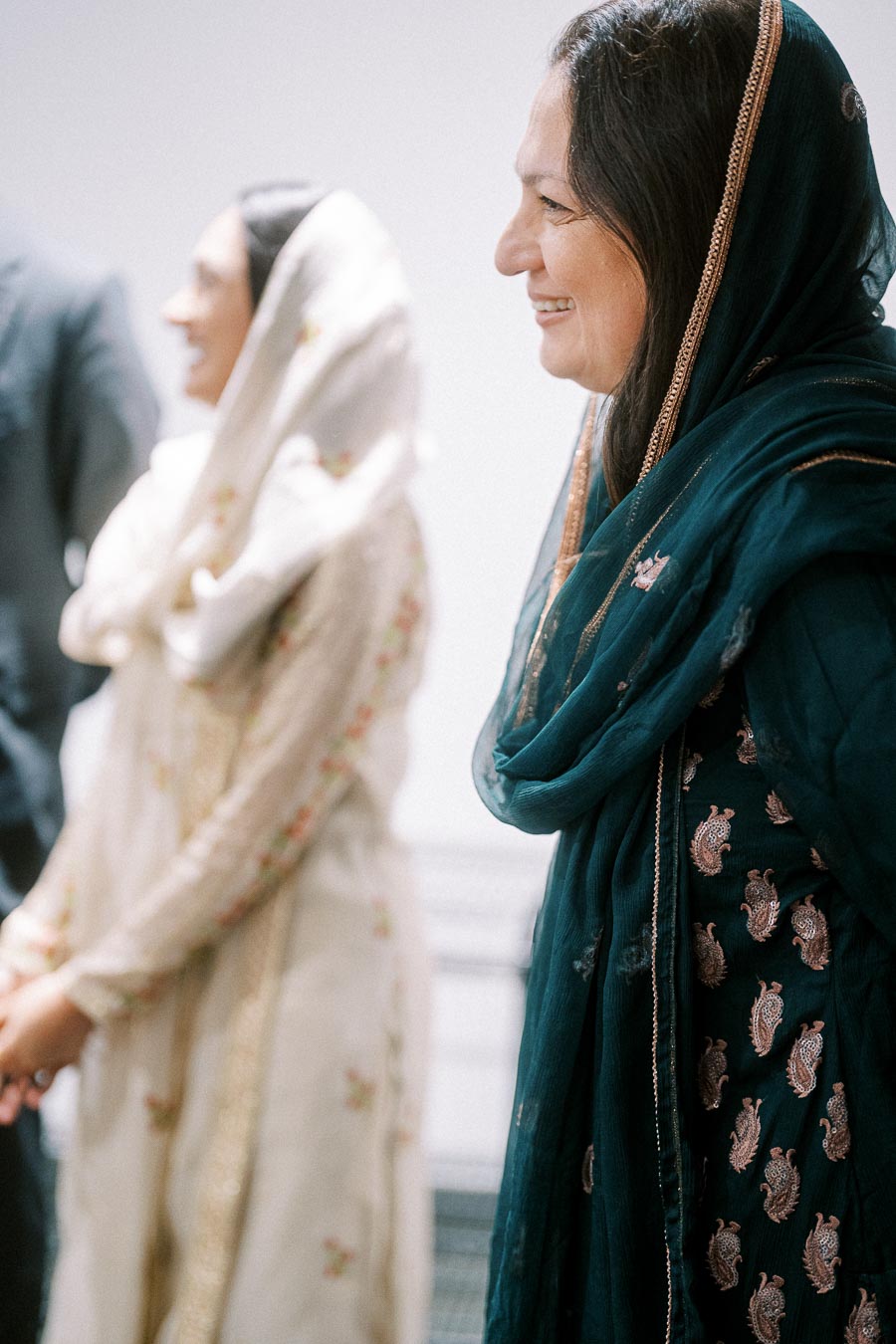 Two women in traditional South Asian attire, one wearing a blue embroidered shawl and the other in a white floral outfit, smiling indoors.