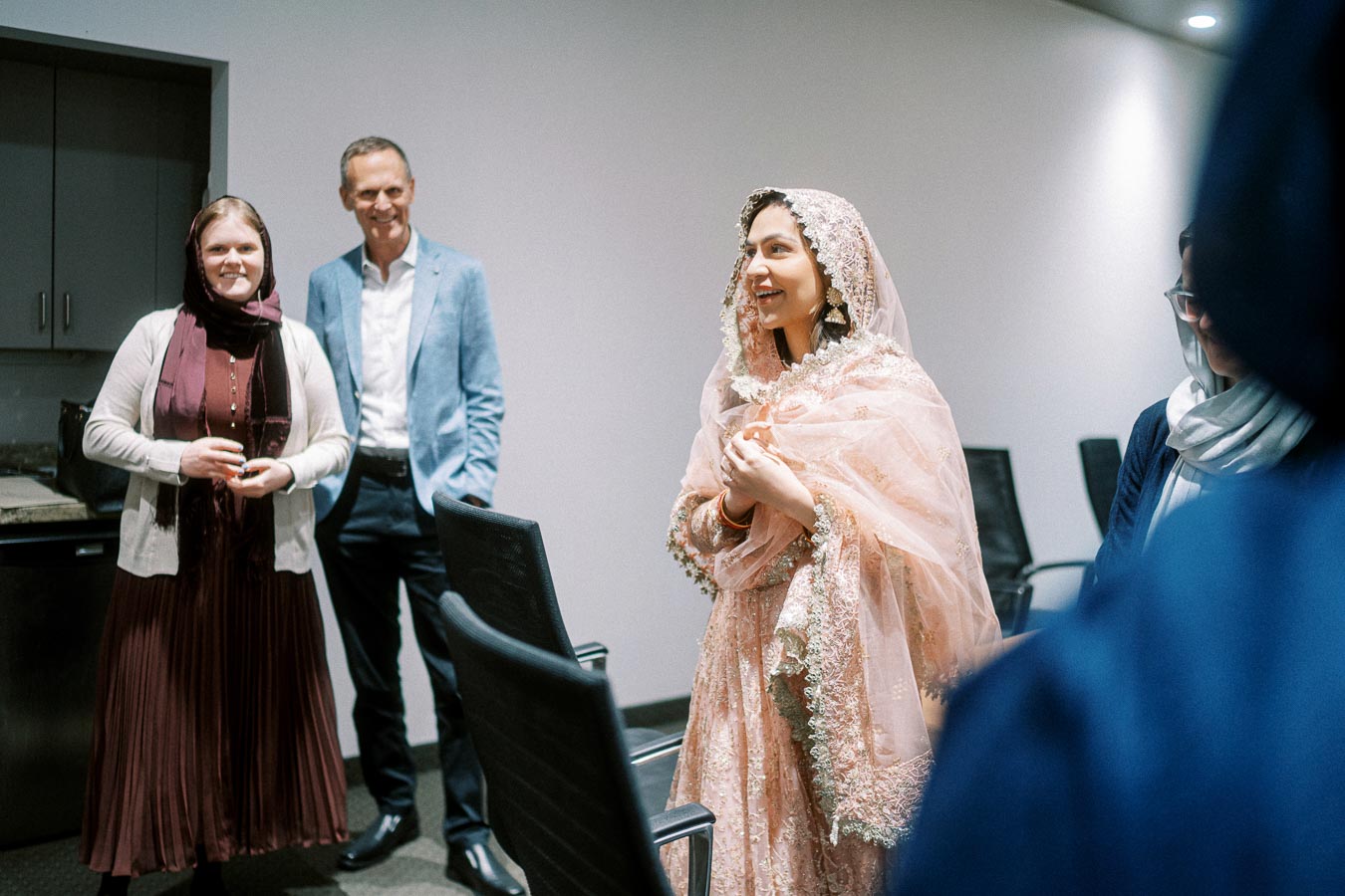 A woman in an elegant, traditional outfit with intricate lace details is smiling and interacting with a group of people in a modern conference room setting. She is surrounded by a man in a blue blazer and a woman in a burgundy dress.