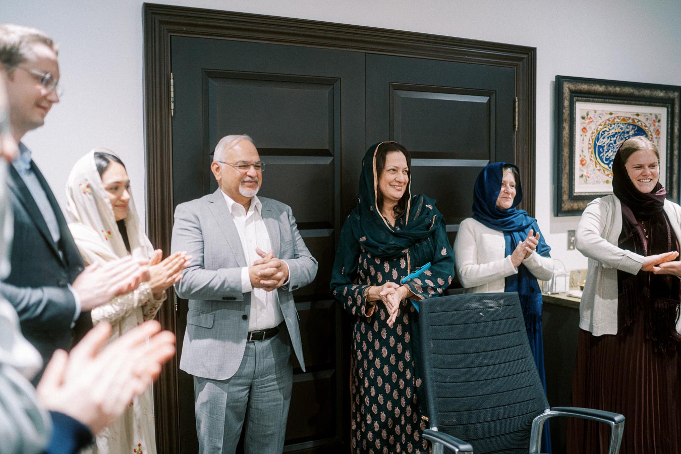 Group of people applauding in a formal meeting room, with diverse attire including suits and traditional clothing, standing in front of dark wooden doors and a framed artwork on the wall.