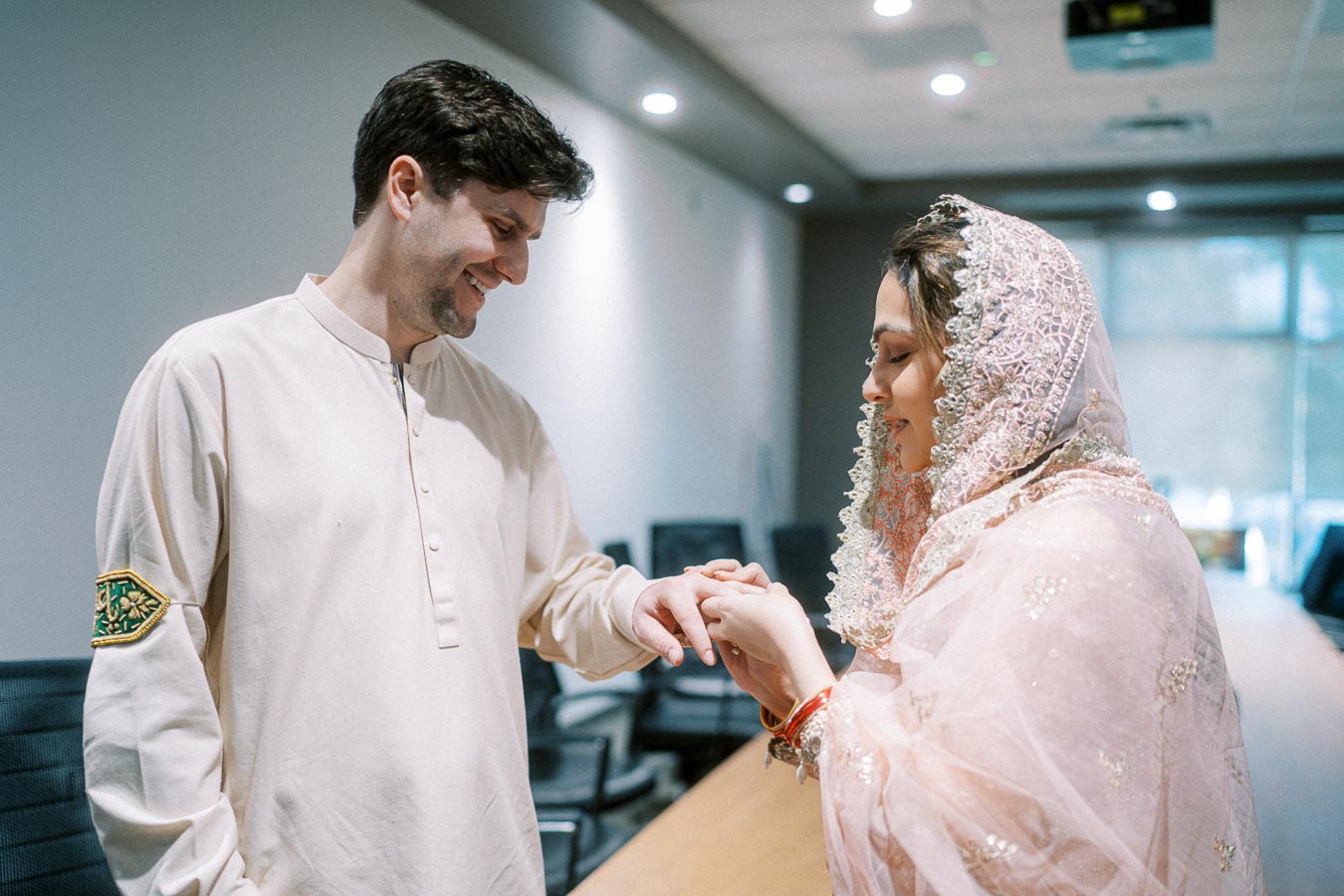 A couple in traditional attire smiling during an engagement ceremony, with the woman placing a ring on the man's finger in a conference room setting.