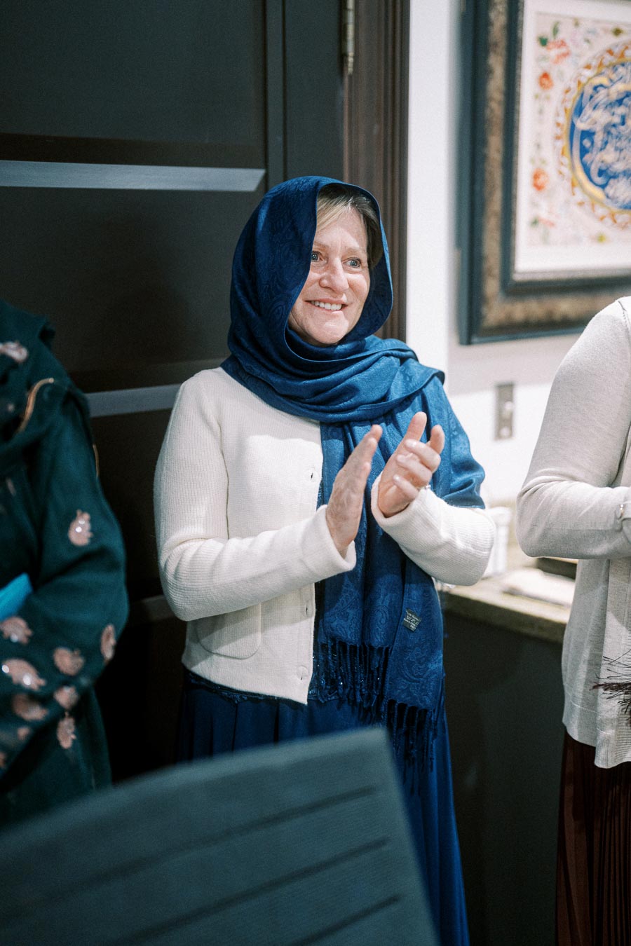 A person smiling and clapping, wearing a blue headscarf and white sweater indoors, with framed artwork on the wall in the background.