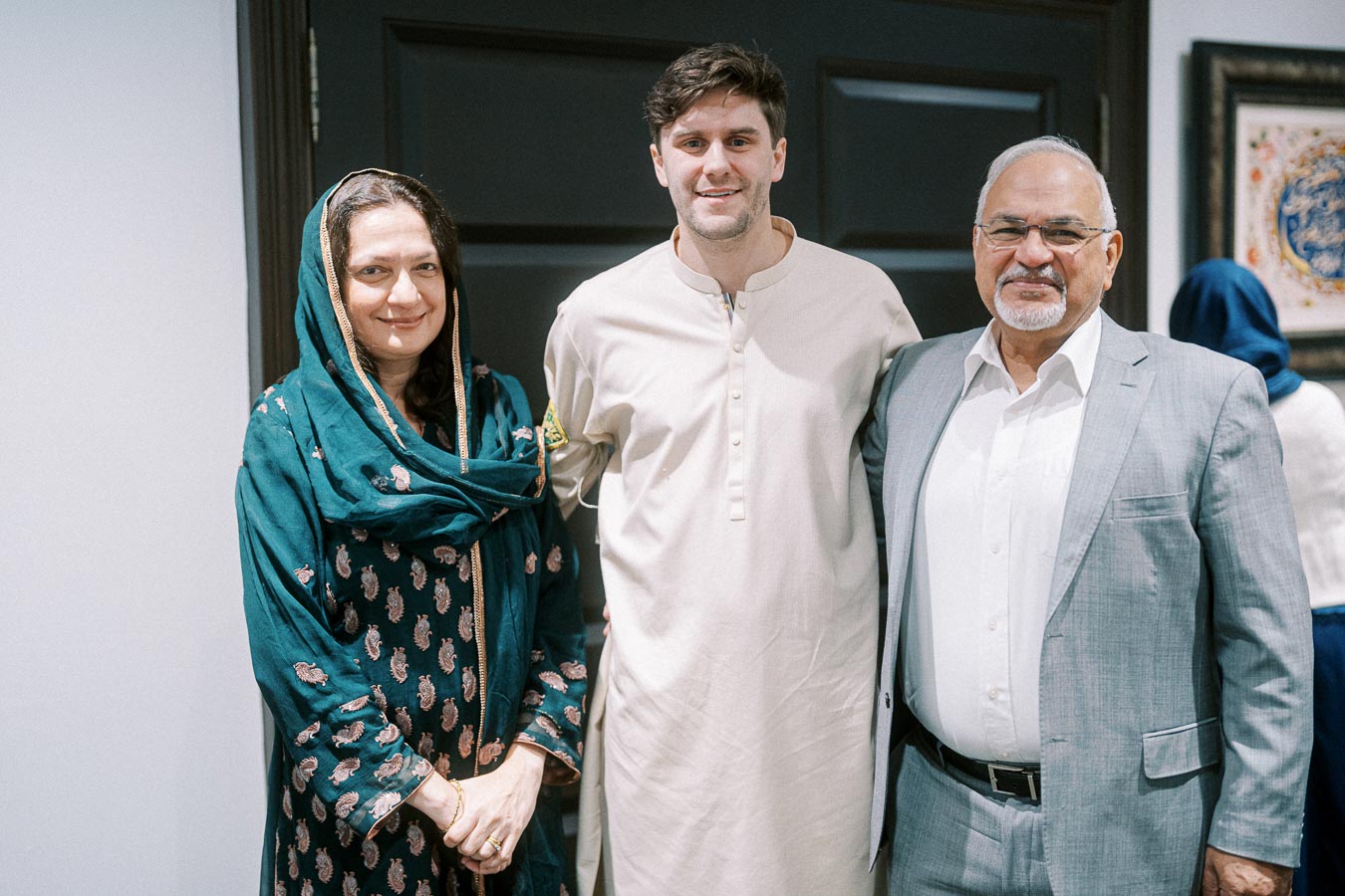 A diverse group of three people smiling indoors, with one person wearing traditional South Asian attire and another in a light suit. They stand in front of a dark door with artwork visible in the background.