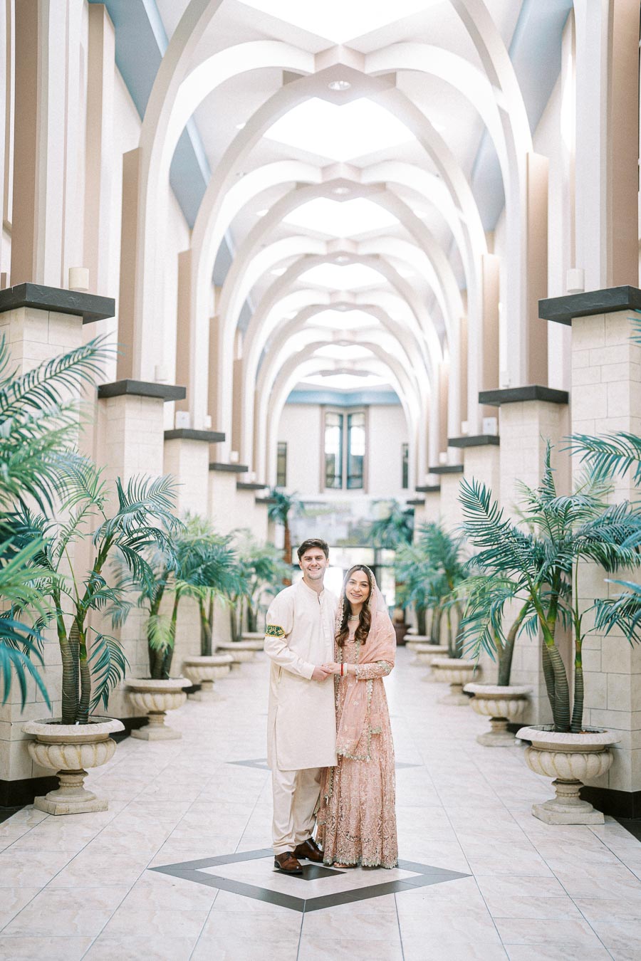 A couple in traditional attire poses in a grand hallway with high, arching ceilings and lush green plants in ornate planters.