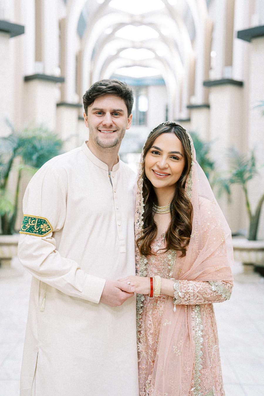 Couple dressed in traditional attire during a wedding celebration, standing in an elegant architectural setting with intricate columns and greenery.