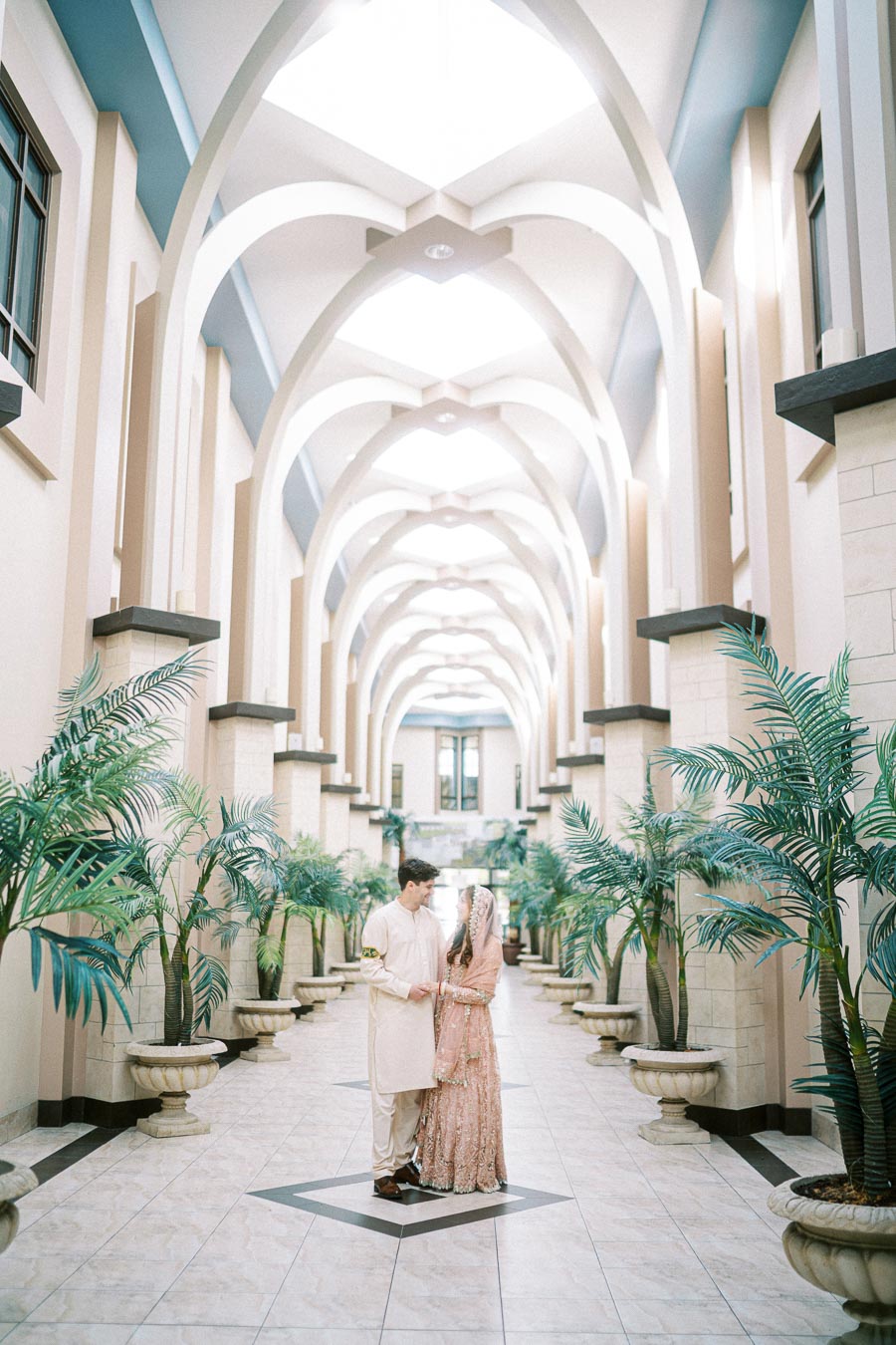 A couple in traditional cultural attire stands in an ornate, arched hallway adorned with potted plants, creating an elegant and serene atmosphere.