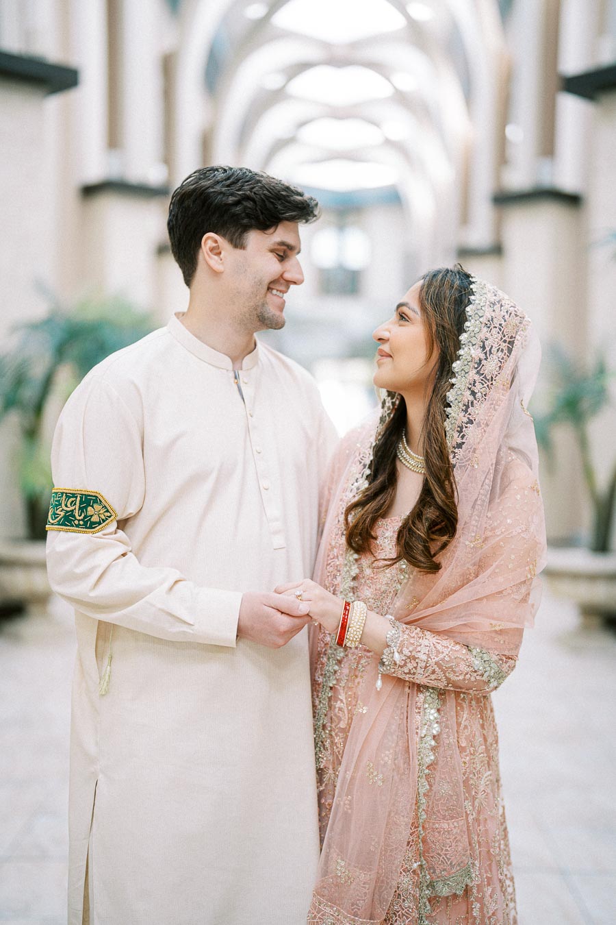Couple smiling at each other in traditional attire, standing in an elegant hallway, showcasing intricate embroidery and cultural fashion.