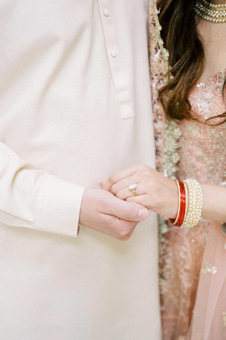 Close-up of couple holding hands, woman wearing traditional South Asian attire with red and pearl bangles and diamond ring, showcasing cultural wedding jewelry and clothing.