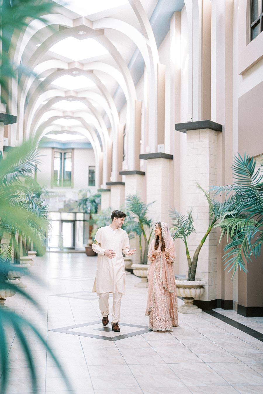 Couple in traditional attire walking in a beautifully decorated hallway with arched ceilings and potted plants.