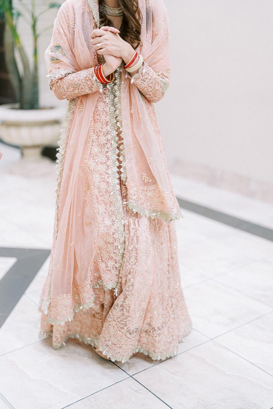 Woman in an elegant, embroidered pink traditional dress with intricate details, red bangles, and delicate veil, standing indoors with a blurred background.