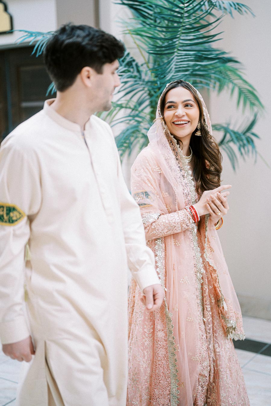 A woman in an elegant pink traditional outfit smiles at a man in a cream-colored kurta, set against a backdrop of lush green foliage, capturing a moment of cultural celebration and joy.