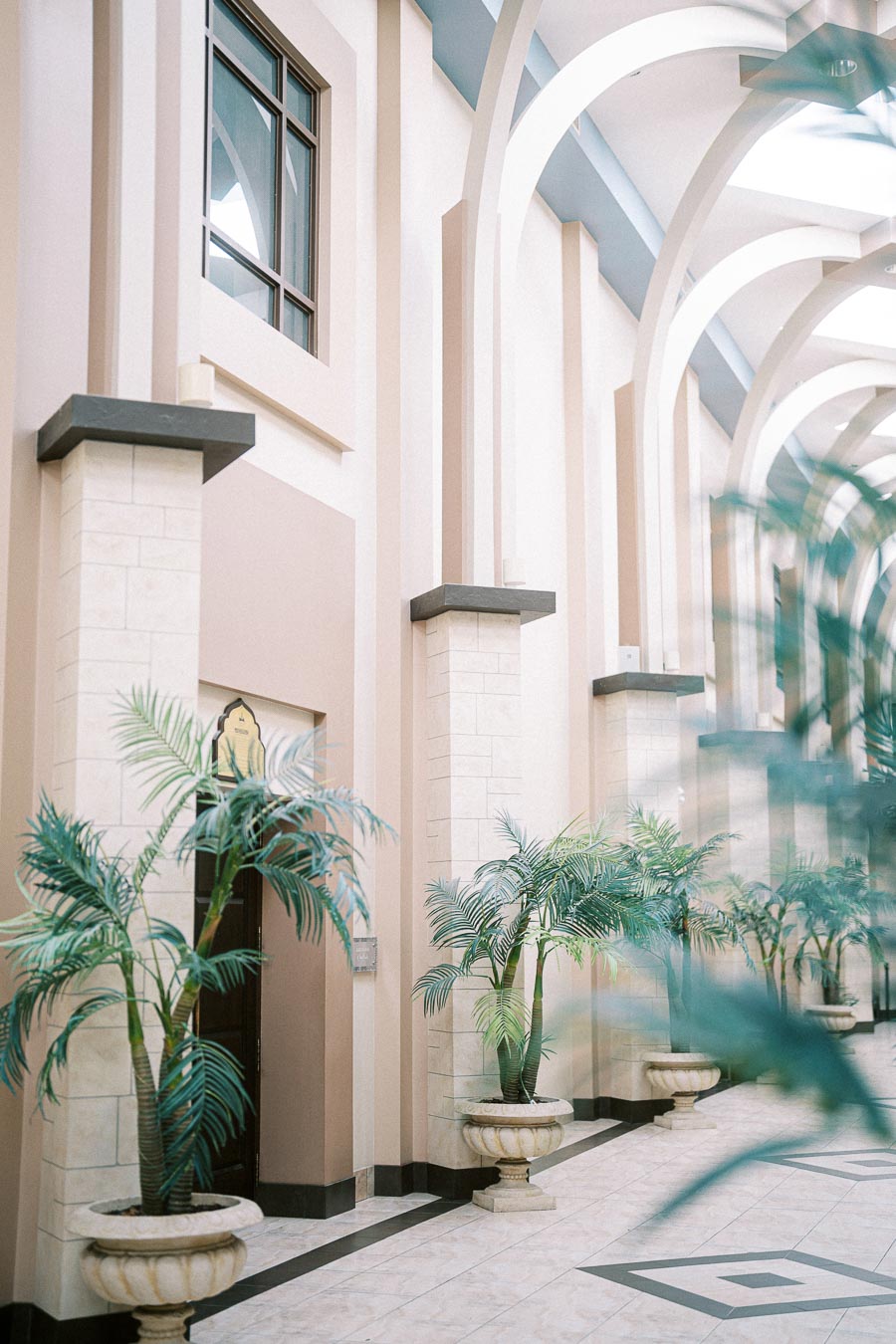 Elegant indoor hallway with tall arches, decorative columns, and potted palm plants lining a tiled walkway.