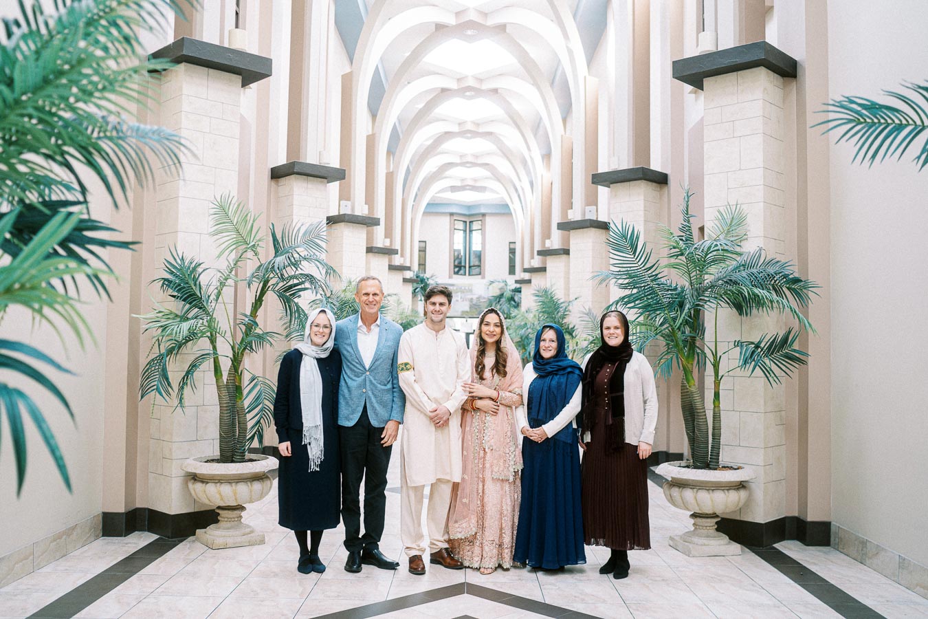 A group of six people stands in a beautifully arched hallway with elegant architectural details and lush greenery. The individuals are dressed in formal attire, with women wearing traditional dresses and headscarves. The bright ambiance and symmetrical design create a serene and sophisticated atmosphere.