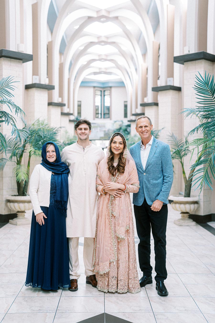 A group photo of four people standing in an elegant indoor hall with high archways and potted plants. The woman on the left wears a blue dress and scarf, the man next to her is dressed in traditional attire, the woman beside him wears a pink embroidered dress, and the man on the right is in a blue blazer.