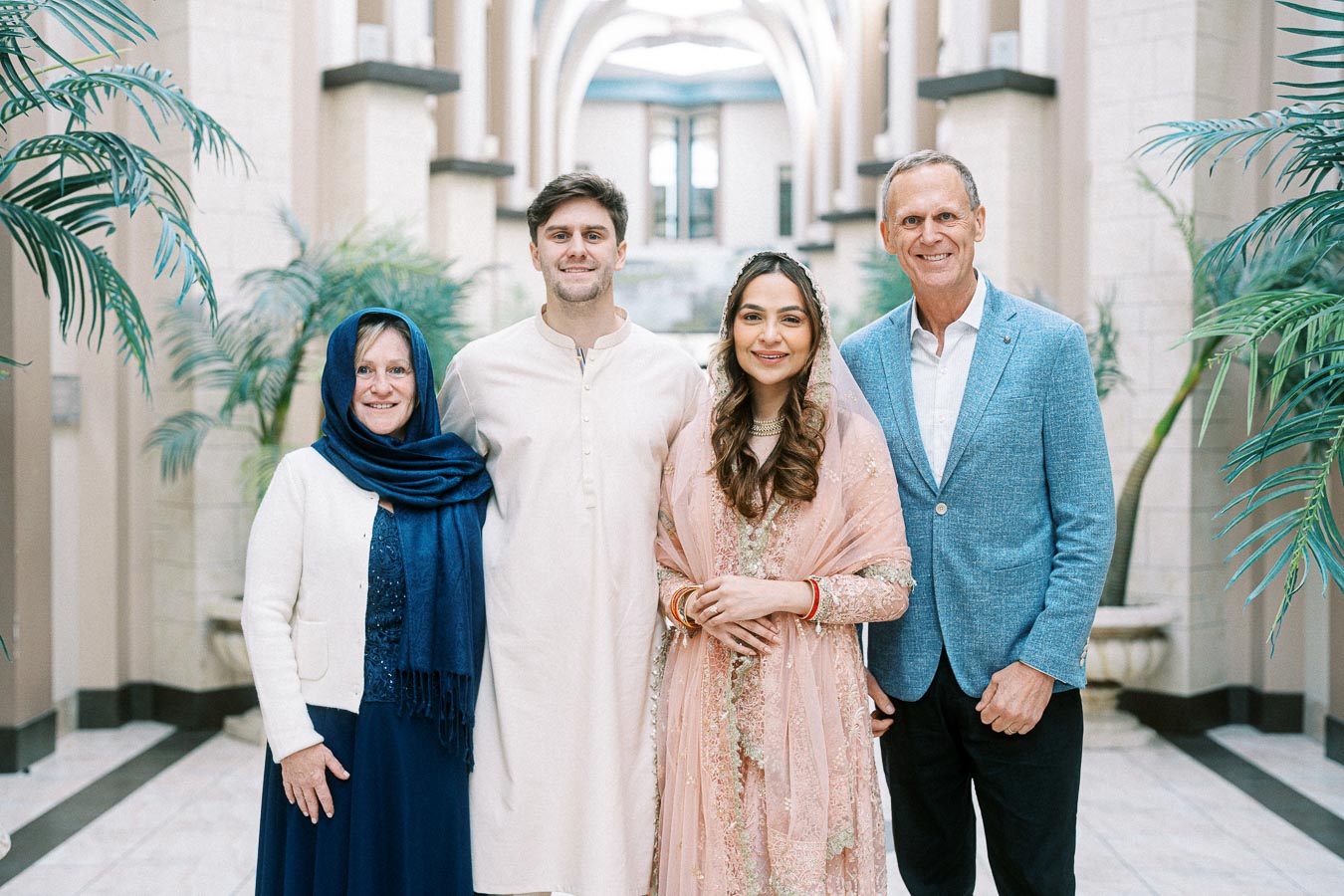 Family portrait featuring four people in traditional and modern attire standing in a beautifully decorated hallway with lush plants.