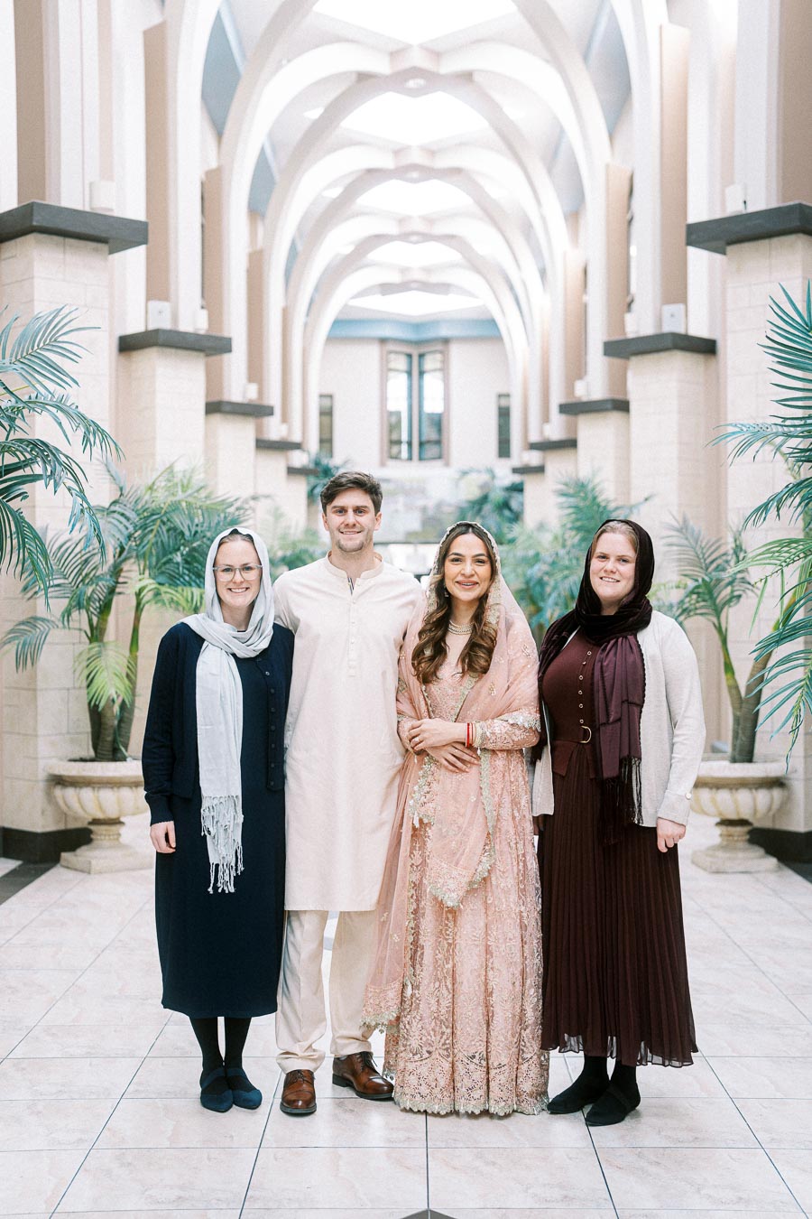Four people posing in an elegant hallway with archways, two women wearing traditional attire, and two women in contemporary outfits, surrounded by lush indoor plants.