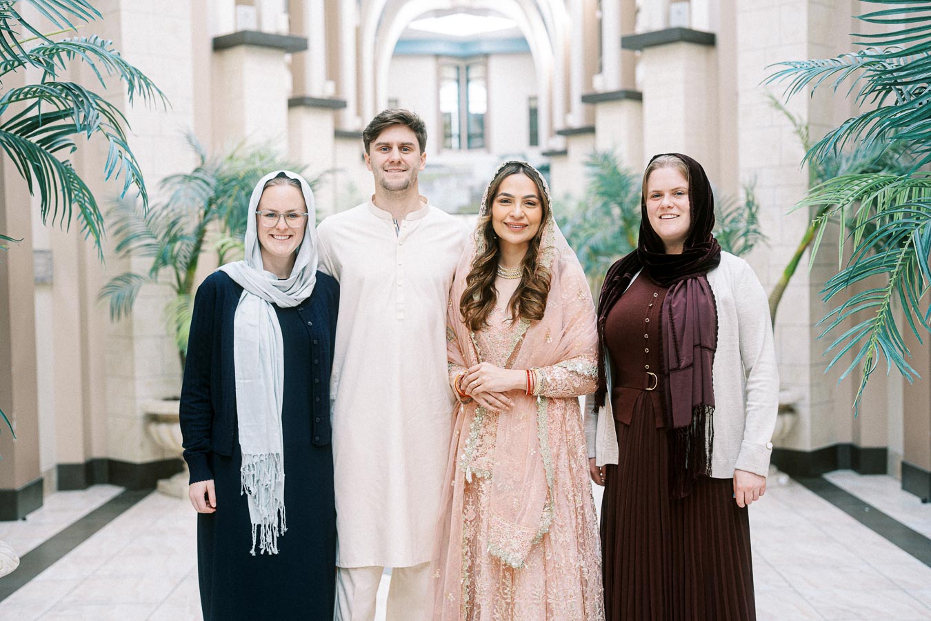 Group portrait of four people dressed in traditional and modern attire, standing in an elegant, arcaded hallway with lush greenery.