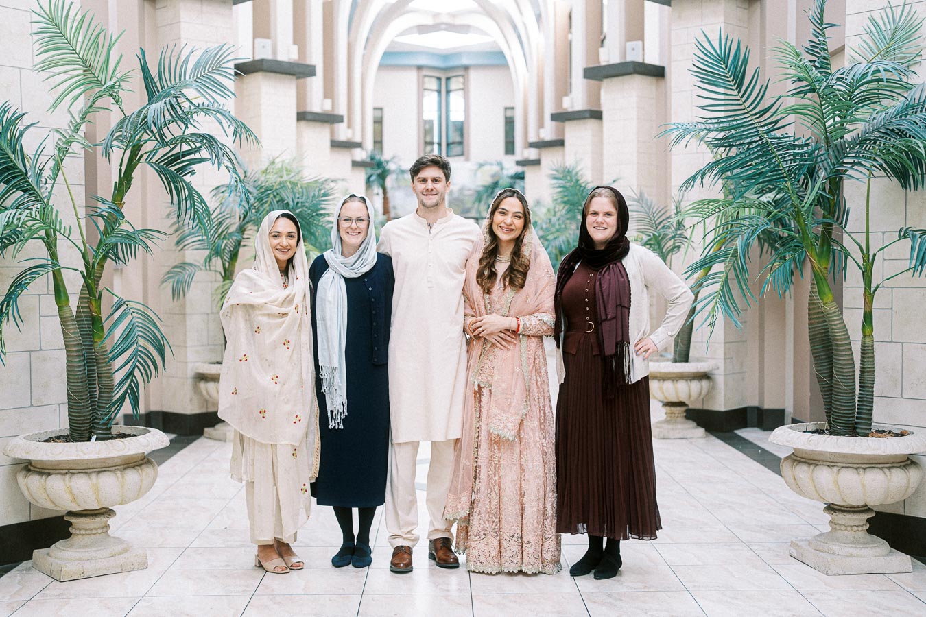Group of people posing in traditional and contemporary attire in an elegant indoor setting adorned with potted palm trees and decorative columns.